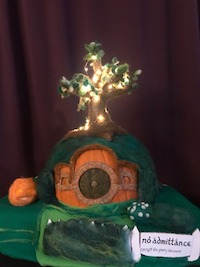A pumpkin covered in felt and felting wool to depict Bag End. The stem has been turned into a tree that is lit up with lights. There is a round door colored on the front and a sign that reads 'No Admittance Except on Party Business' in the front next to a mossy fence. The garden has small green shrubs and a bush covered in white flowers. There is also a small needle felted jack o lantern in the garden. There is a purple curtain in the background.